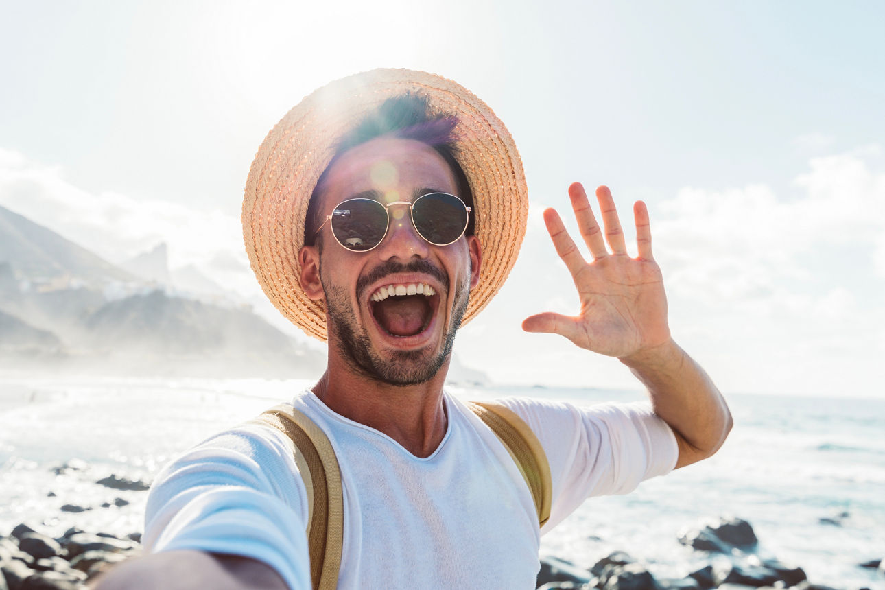 Ein junger Erwachsener wingt lachend in die Kamera während er am Strand steht