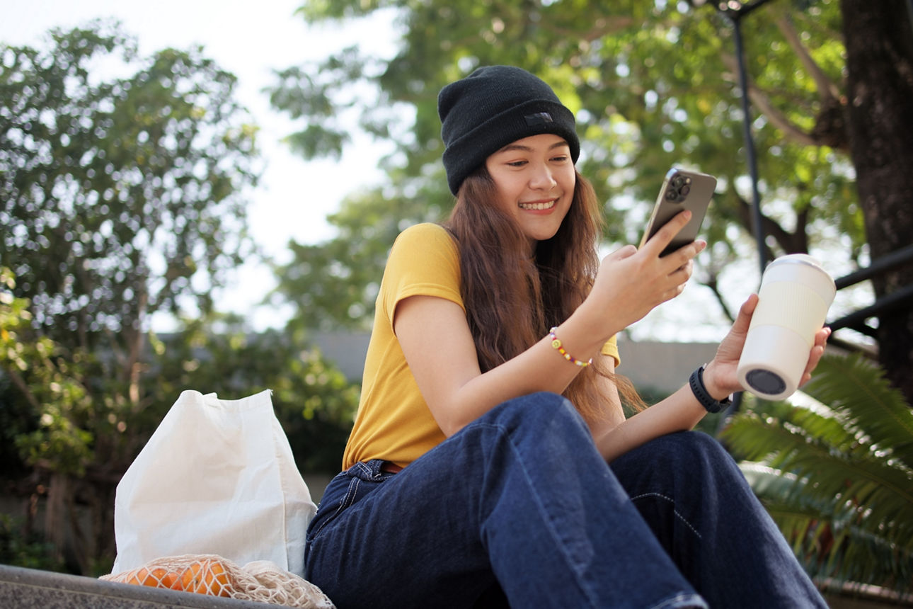 Jugendliche sitzt im Park mit einem Kaffee und ihrem Handy in der Hand