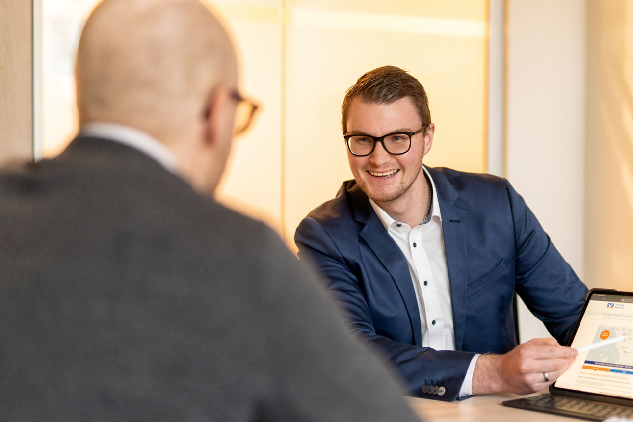 Firmenkundenbetreuer sitzt mit Kundem im Büro und zeigt auf ein Tablet