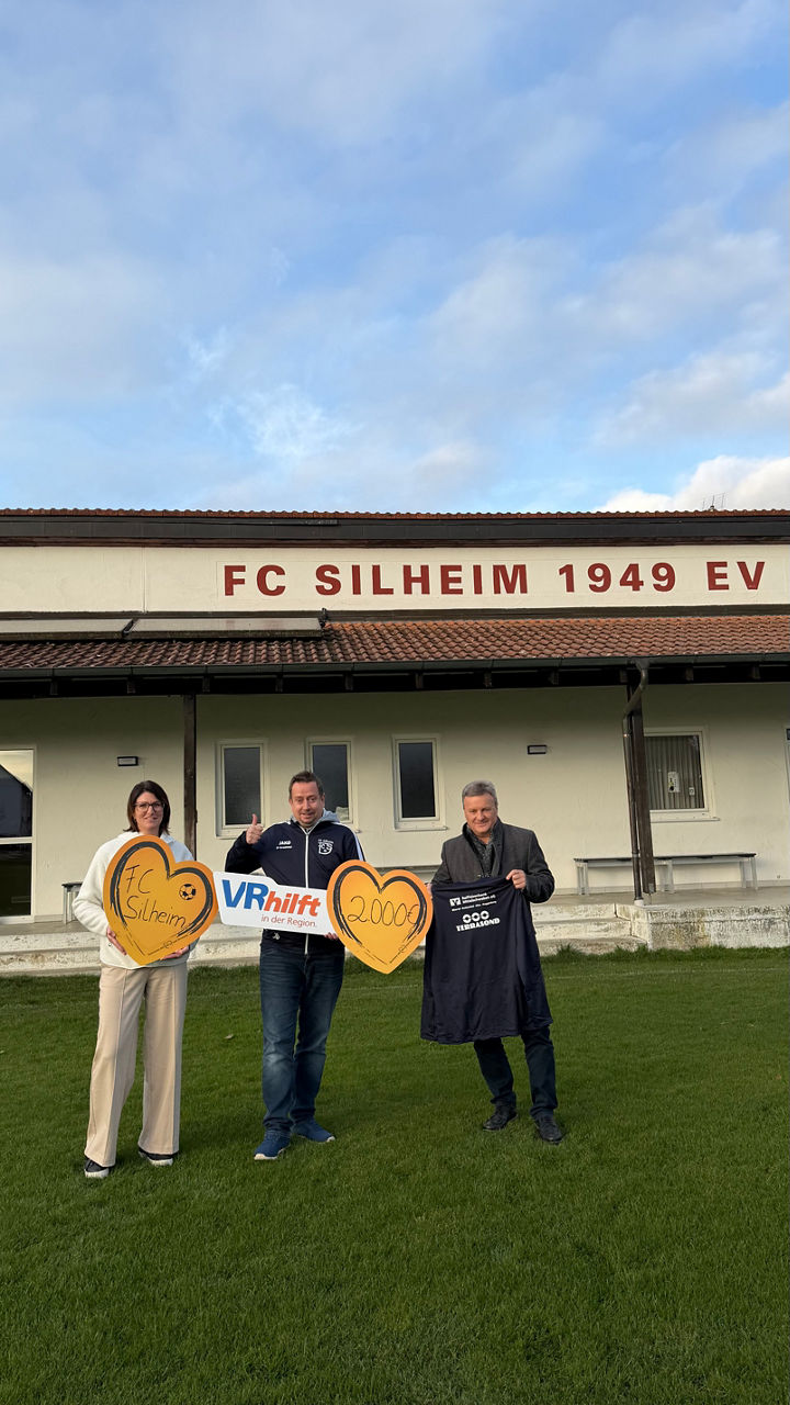 Drei Personen vor Vereinsheim FC Silheim mit Herzschildern und „VR hilft“-Banner