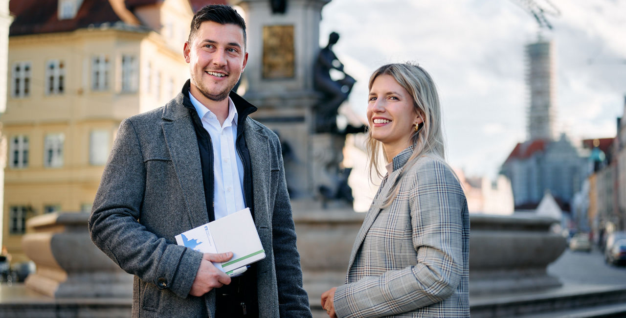 lächelnder Mann und Frau vor Brunnen in Augsburg, beides Mitarbeitende