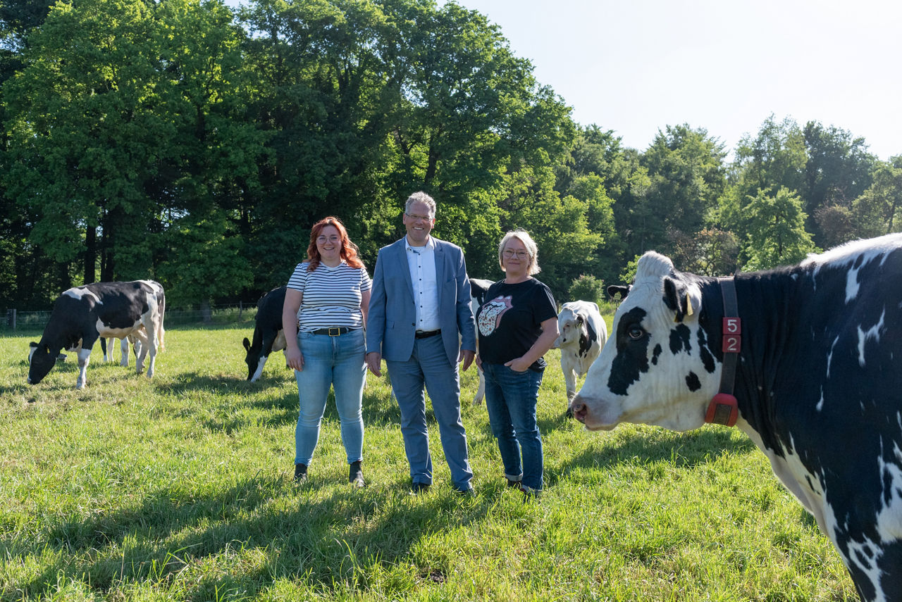 2 Kunden und 1 Firmenkundenberater stehen auf einer landwirtschaftlichen Fläche mit Kühen