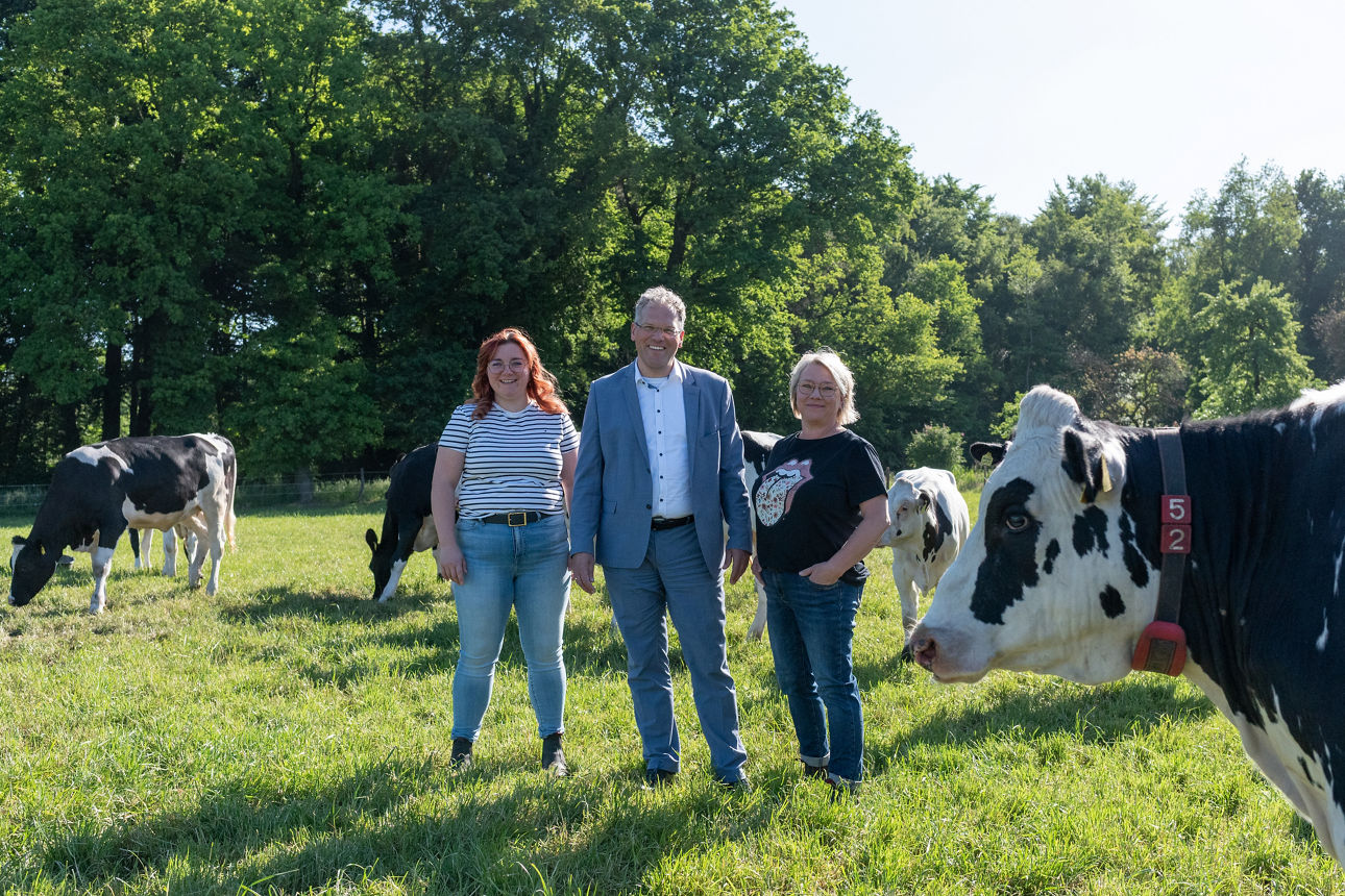 2 Kunden und 1 Firmenkundenberater stehen auf einer landwirtschaftlichen Fläche mit Kühen
