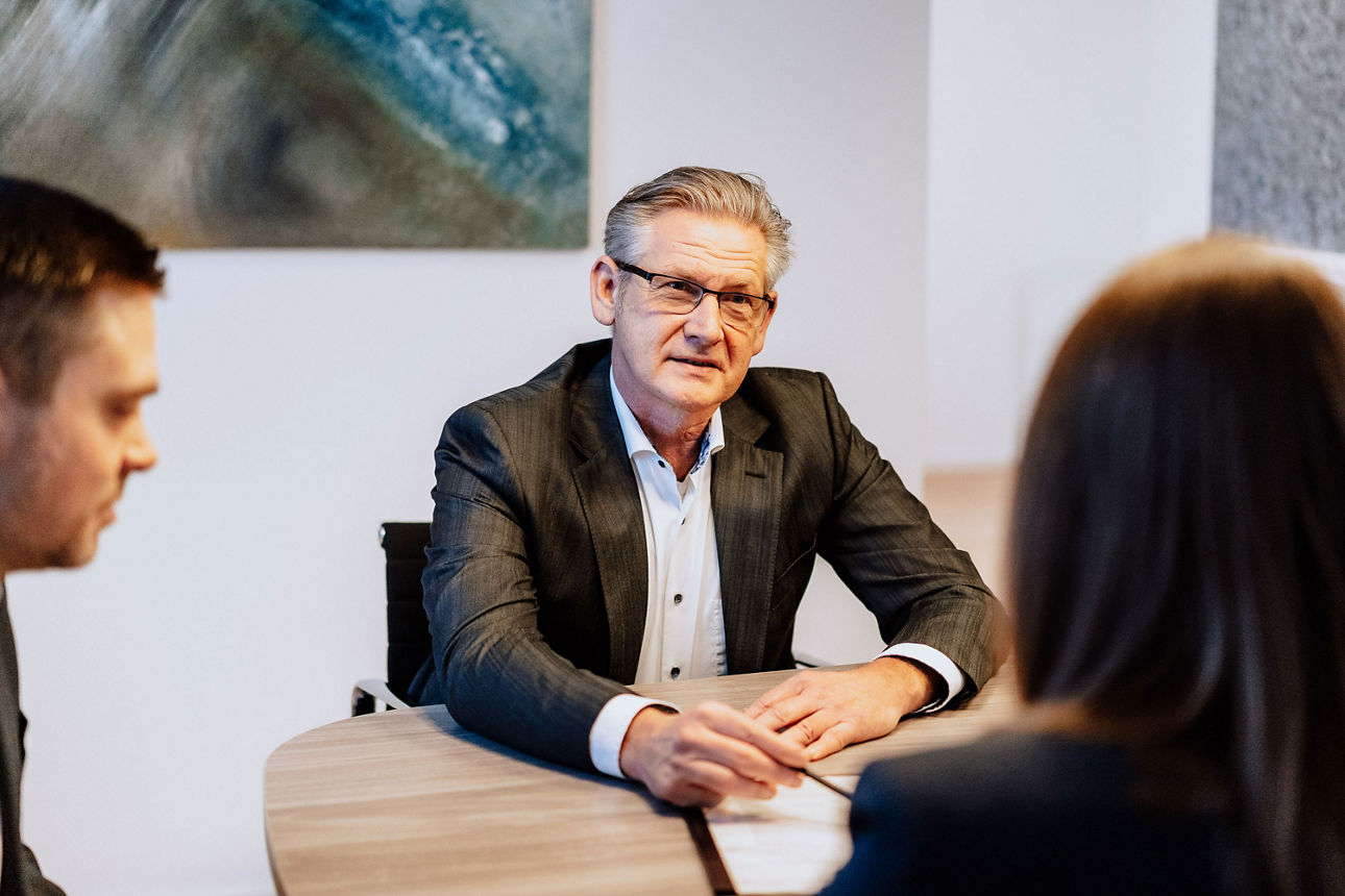 Mann mit grauen Haaren und Brille mit Unterlagen an einem Tisch