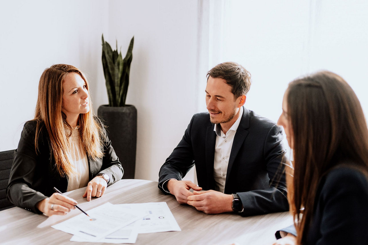 Drei Personen, die sich anschauen, sitzen an einem Tisch mit Unterlagen.