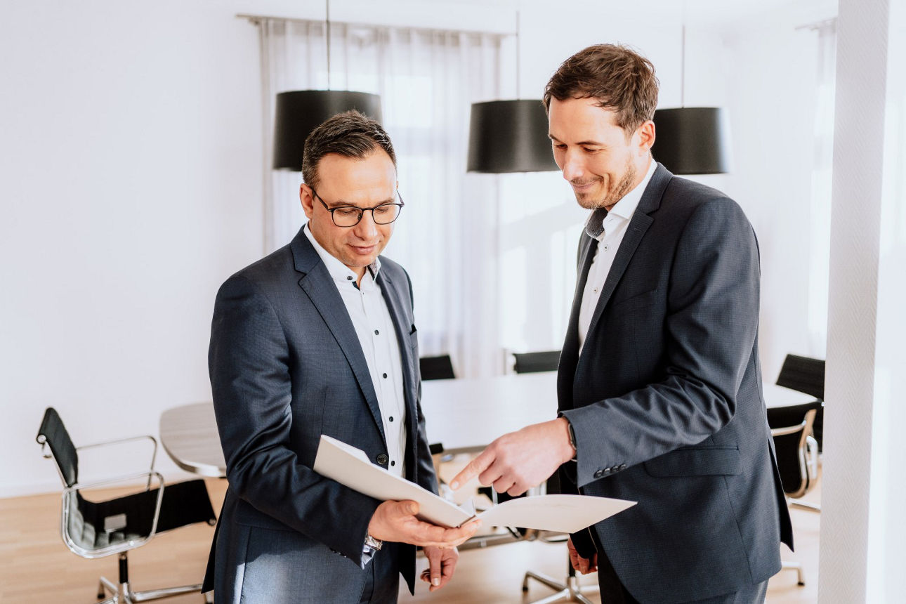 Zwei Männer im Anzug schauen in eine Mappe mit Unterlagen