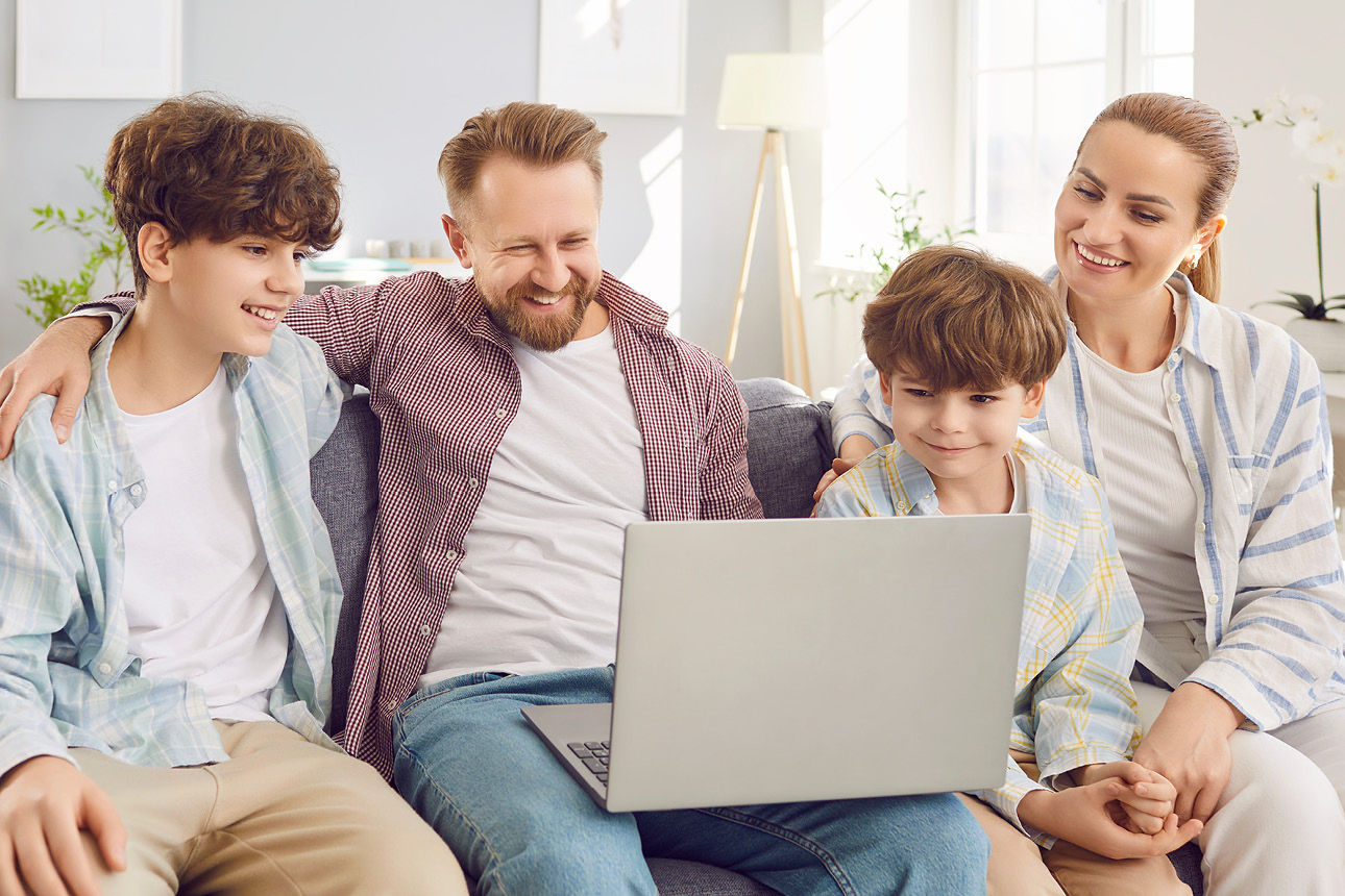 Lächelnde vierköpfige Familie sitzt auf dem Sofa und schaut gemeinsam auf einen Laptop.