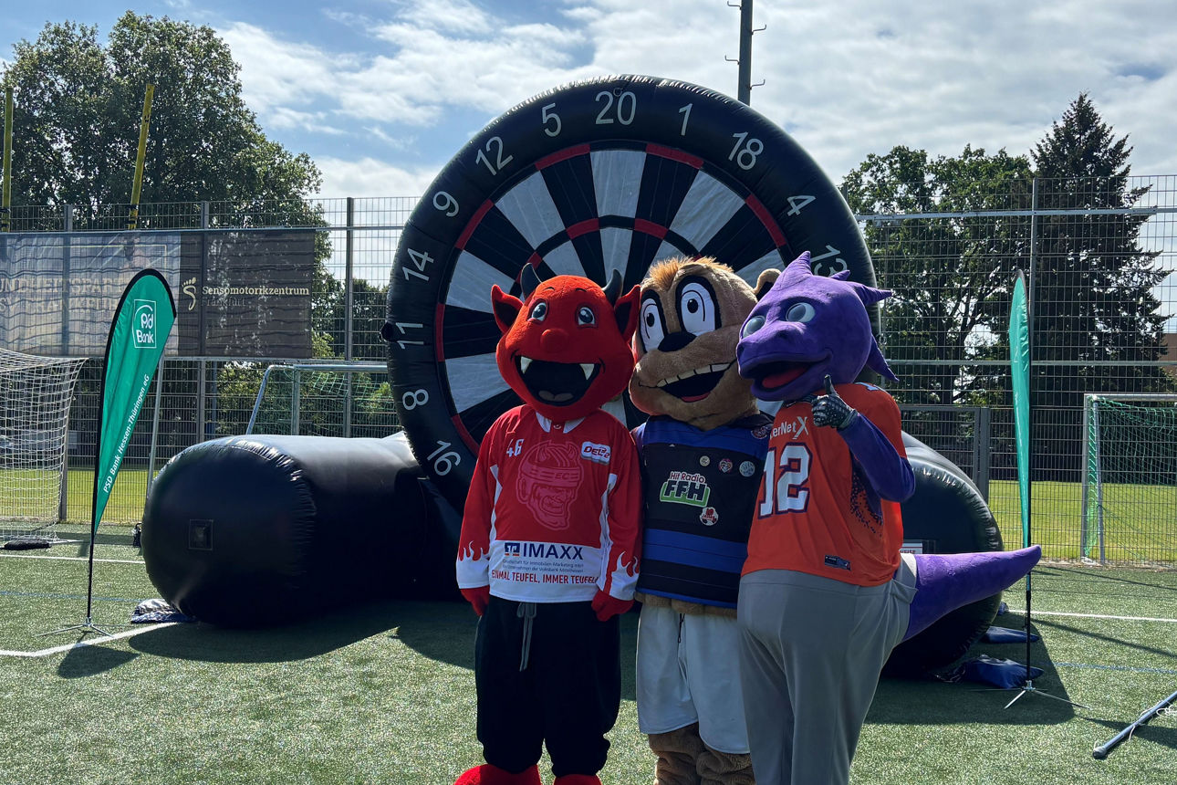 Drei als Maskottchen von Vereinen verkleidete Personen stehen vor einer XXL-Fußball Dartscheibe