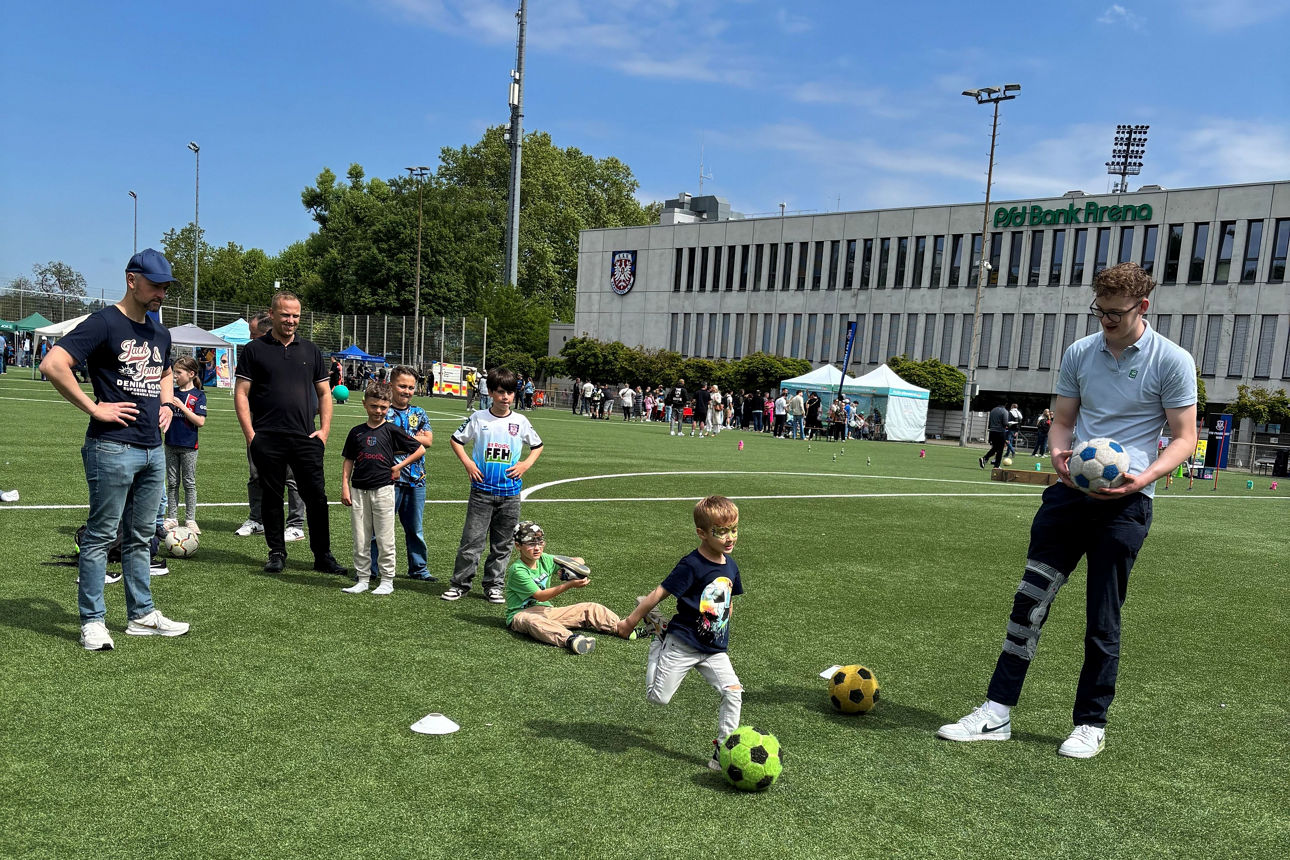 Kinder schießen auf einem Rasenplatz auf einen Fußball, im Hintergrund eine Fußballarena