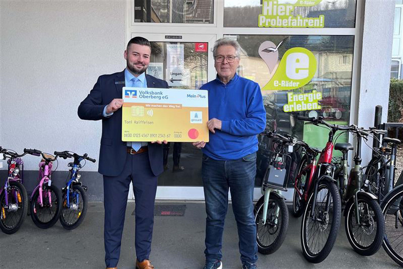 Zwei Männer lächeln in die Kamera und halten gemeinsam eine übergroße, symbolische Bankkarte der Volksbank Oberberg.