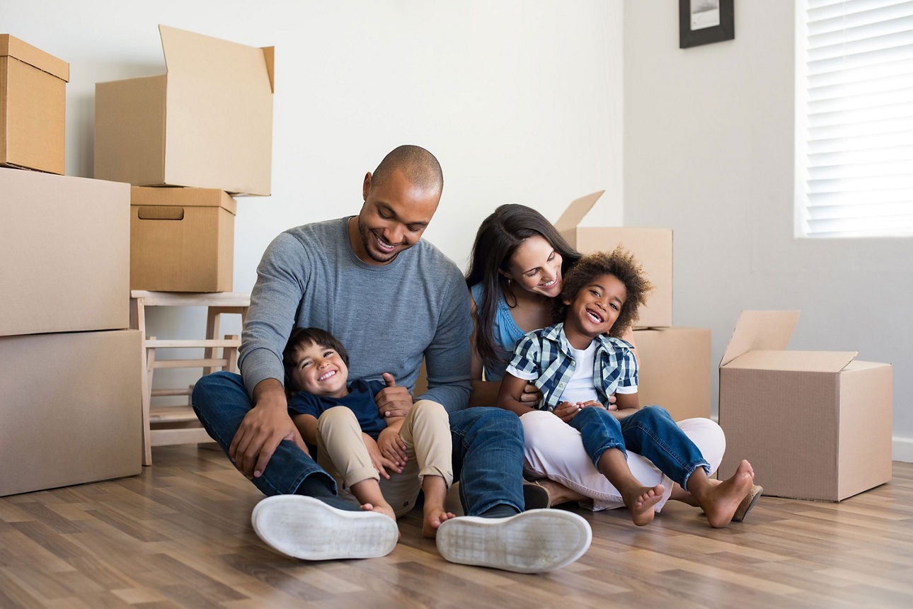 Familie mit Kindern sitzt im Zimmer auf dem Boden. Es stehen viele Umzugskartons im Hintergrund.