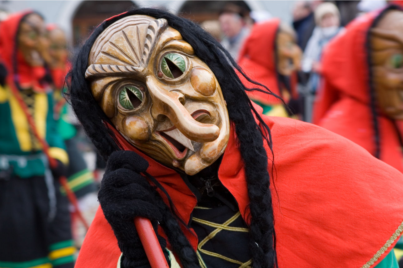 Eine Hexe mit einer Holzmaske und einem roten Umhang auf dem Faschingsumzug.