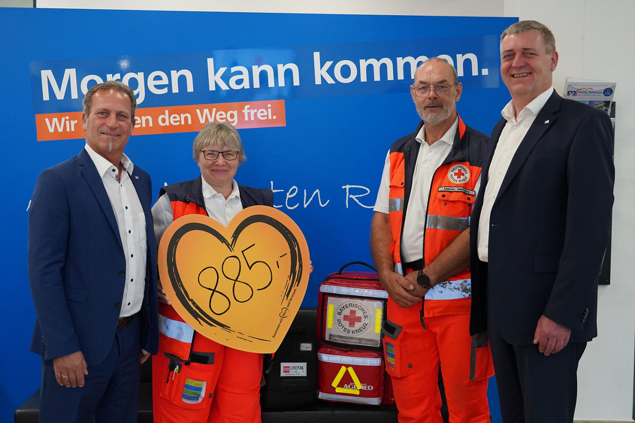 Eine Frau und ein Mann in Rettungsdienstkleidung posieren mit zwei Männern in Anzügen. Sie halten ein Herz in der Hand.