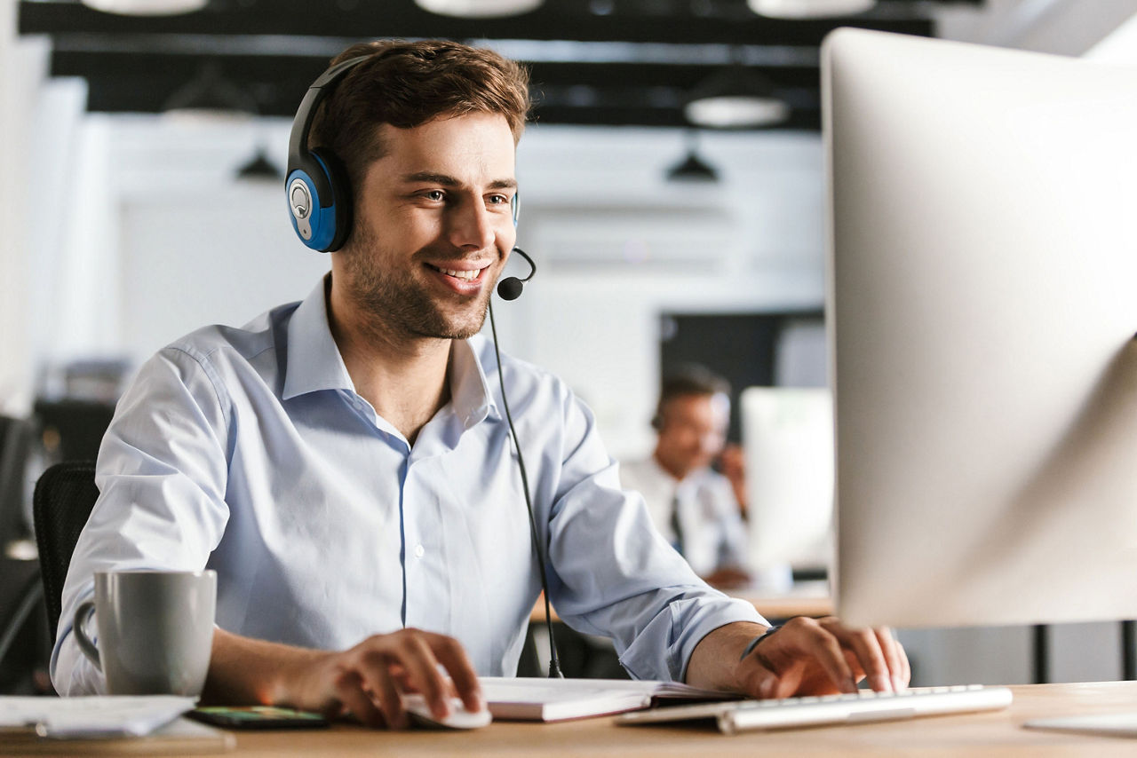 Foto eines jungen Arbeiters 20 jahre in Bürokleidung und Headset, lächelnd und mit Kunden im Callcenter sprechend