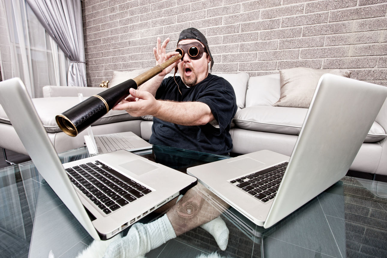Funny photograph of a man with three laptops sitting on the floor of his living room looking at the monitor through a hand-held telescope with a look of surprise on his face. Internet security, computer surveillance, remote network support, and identity theft concepts.