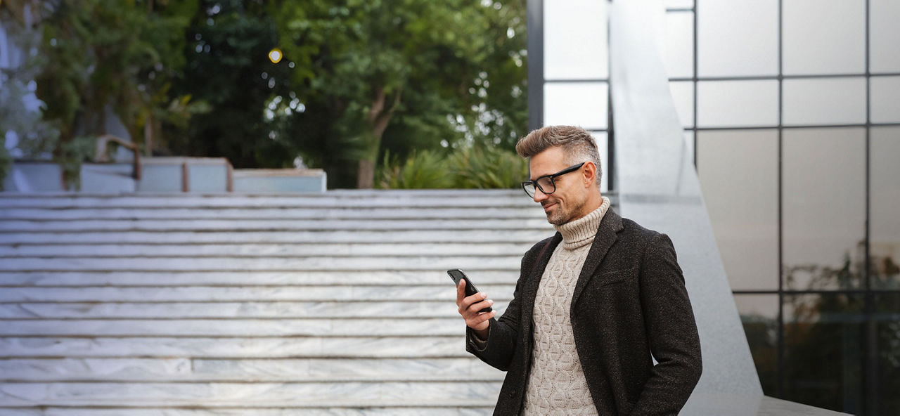 Junger lächelnder Mann mit Brille und einem schwarzen Mantel steht neben einer hellen Steintreppe und  schaut in sein Smartphone