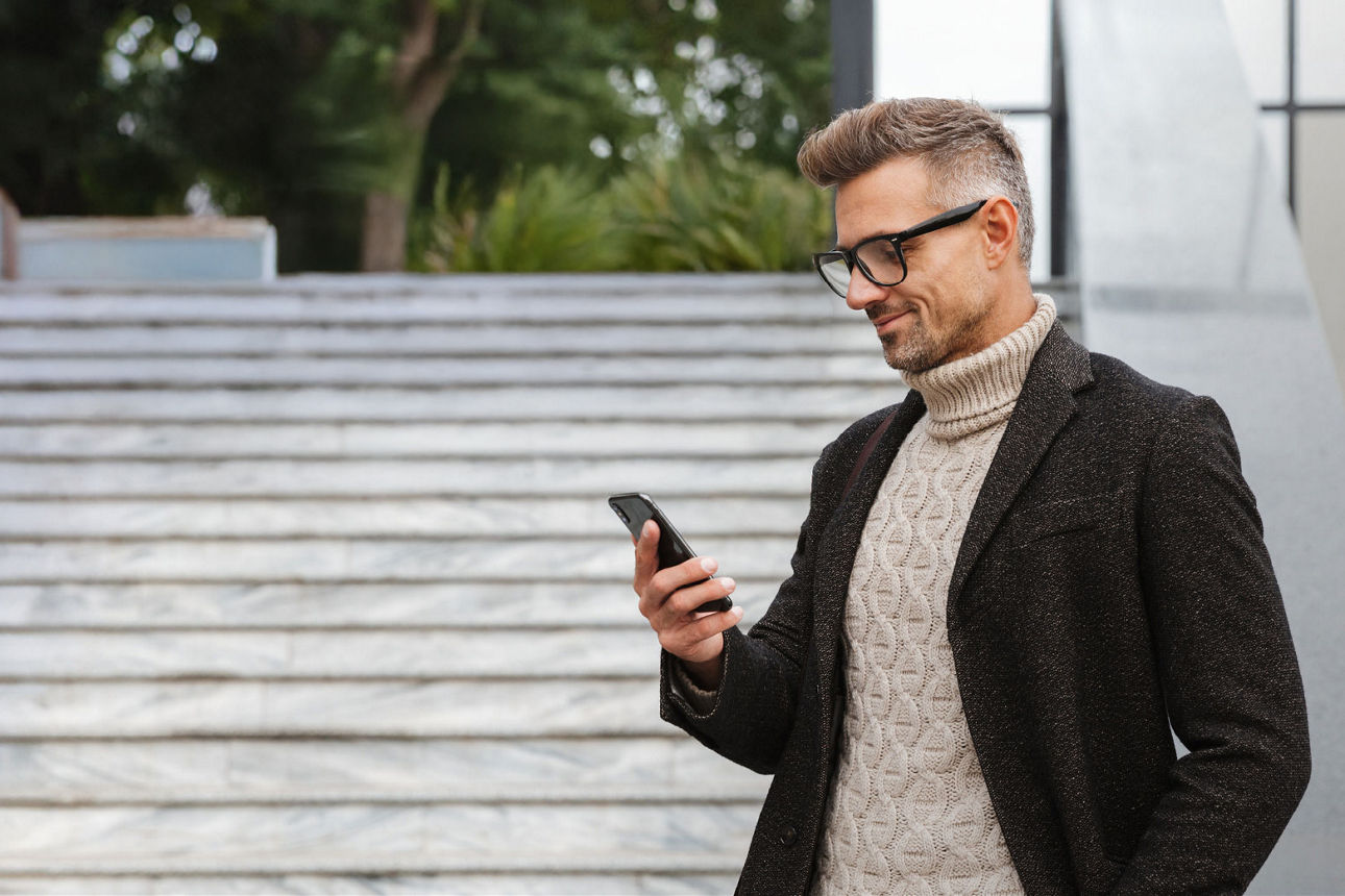 Junger lächelnder Mann mit Brille und einem schwarzen Mantel steht neben einer hellen Steintreppe und  schaut in sein Smartphone