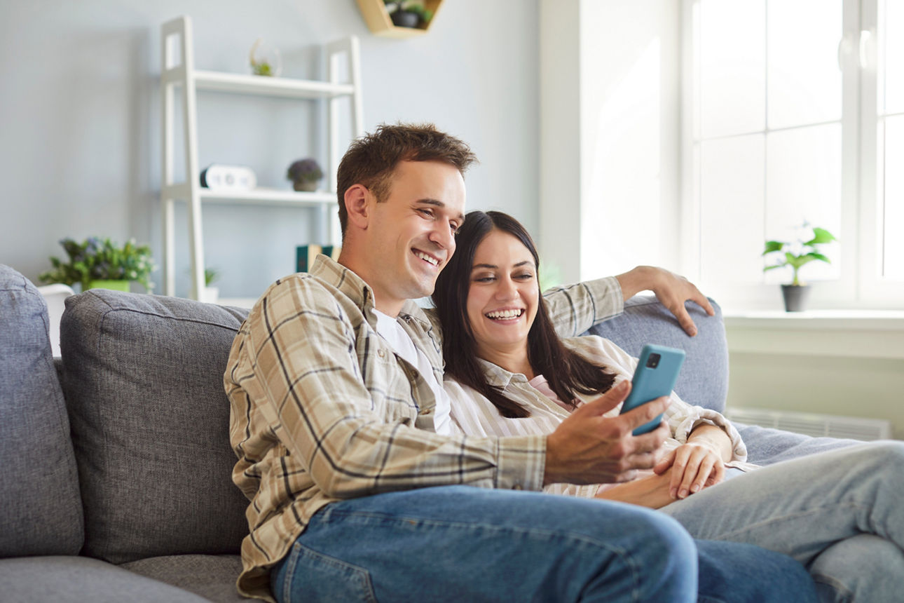 Ein junges glückliches Paar sitz auf dem Sofa und schaut gemeinsam ins Handy.