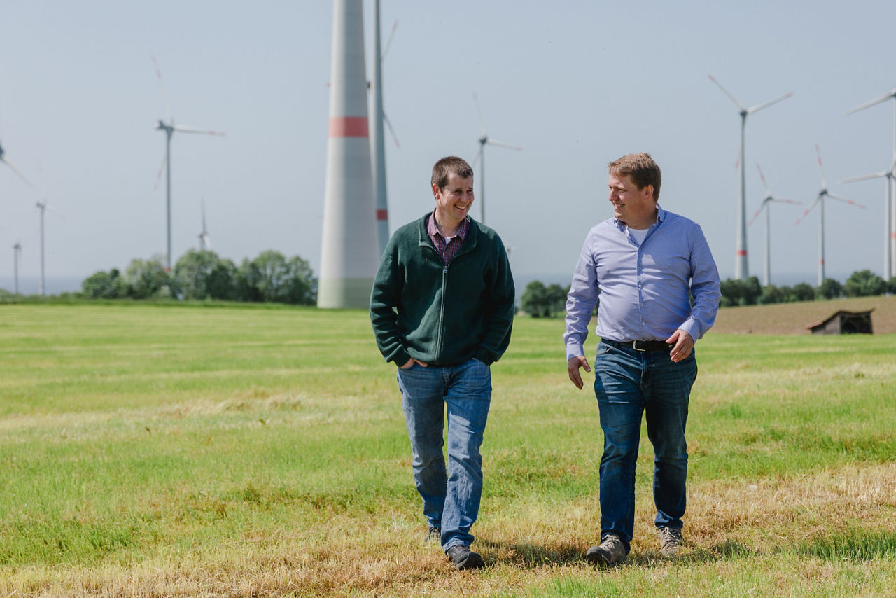 Zwei Personen, die auf einer Wiese laufen. Im Hintergrund sind mehrere Windräder zu sehen.