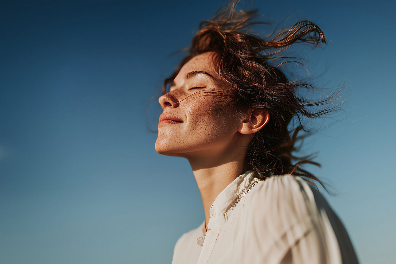 Frau mit braunen, vom Wind zerzausten Haaren, trägt eine weiße Bluse, vor blauem Himmel.