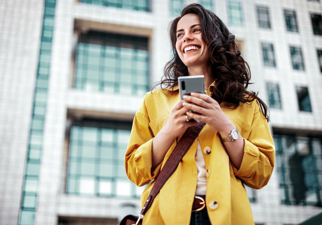 Frau mit Smartphone steht lächelnd vor einem Gebäude