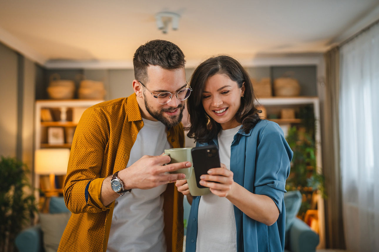 Zwei Personen stehen nah beieinander in einem Wohnzimmer und schauen gemeinsam auf ein Smartphone