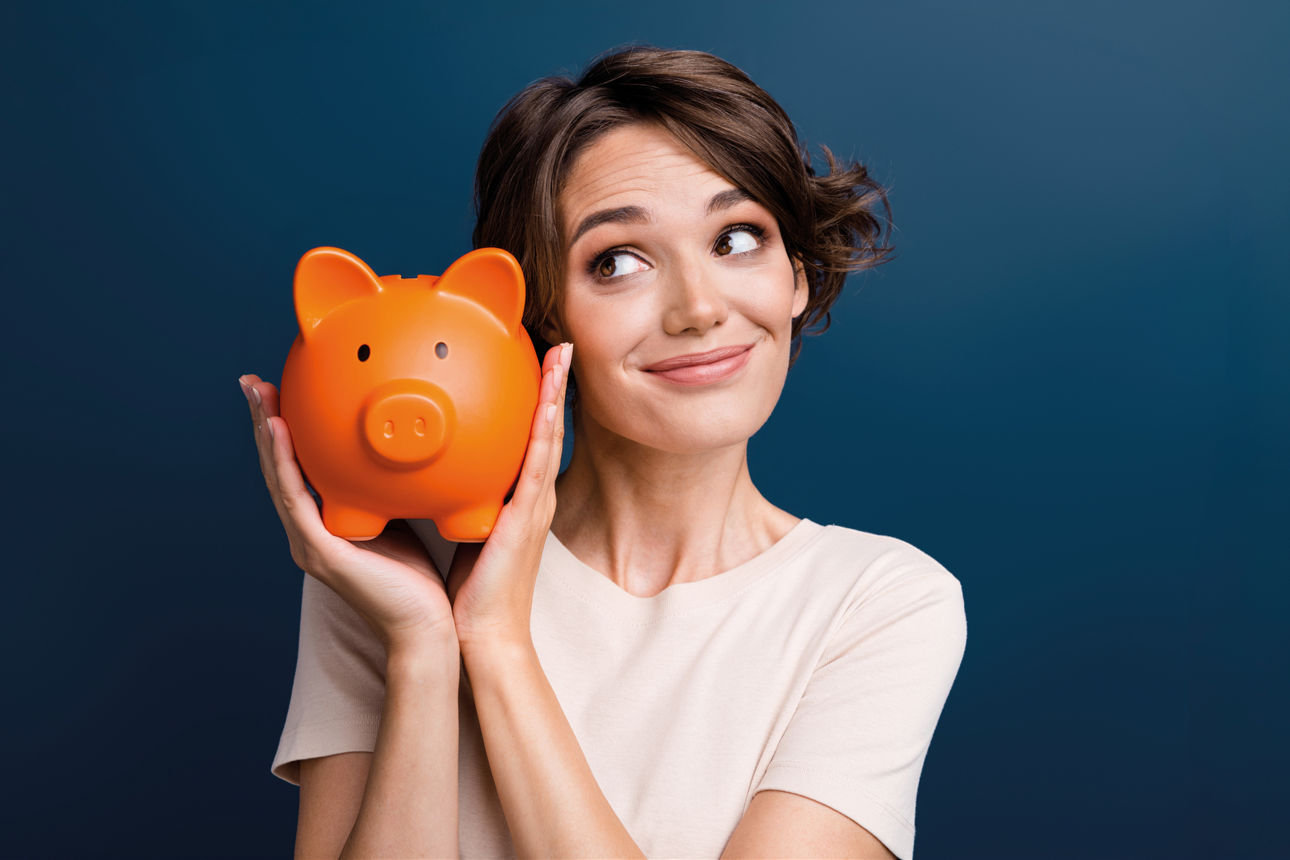 Eine Frau hält ein orangefarbenes Sparschwein mit beiden Händen vor einem dunkelblauen Hintergrund und blickt nach oben rechts. Das Bild symbolisiert Sparen und Vermögensaufbau.