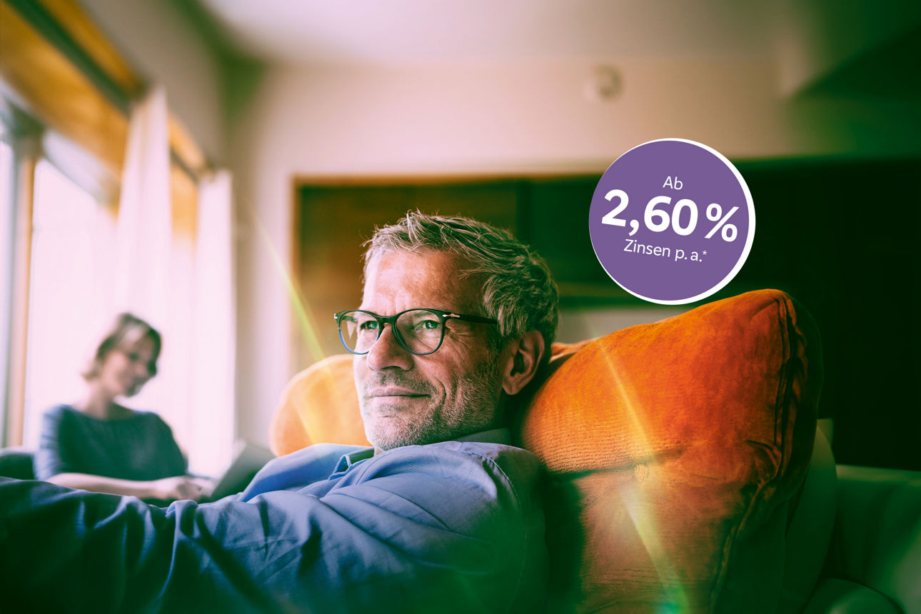 Mann mit Brille sitzt gemütlich auf dem Sofa. Im Hintergrund sieht man noch eine Frau.