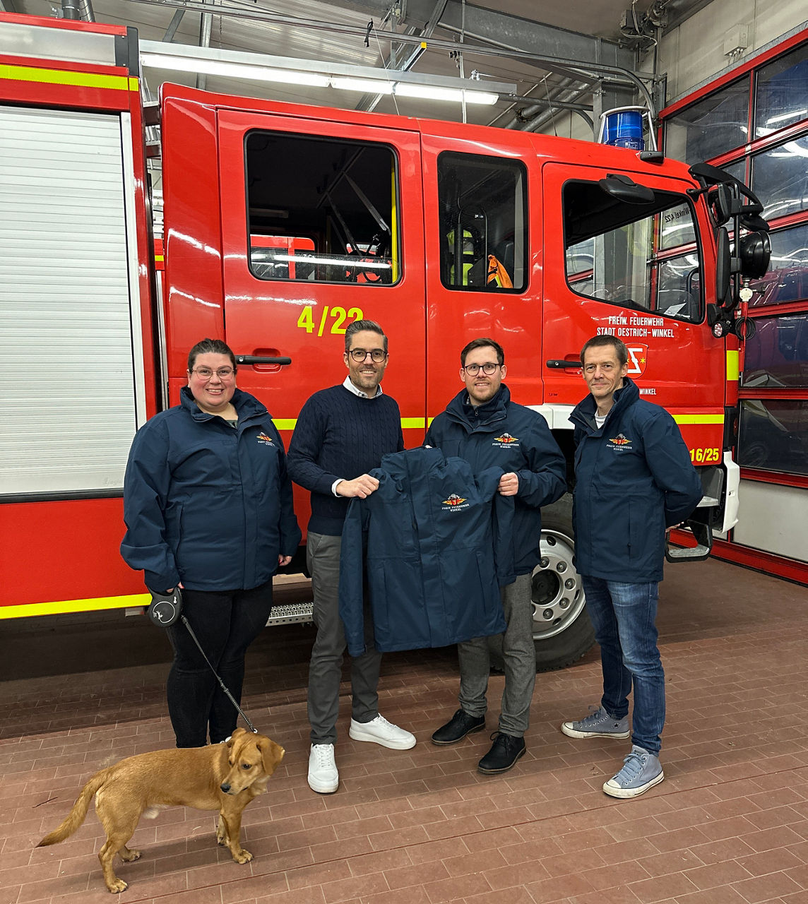 Vier Personen in blauen Jacken stehen vor einem roten Feuerwehrfahrzeug, eine Person hält eine Jacke, ein Hund an Leine steht links im Bild