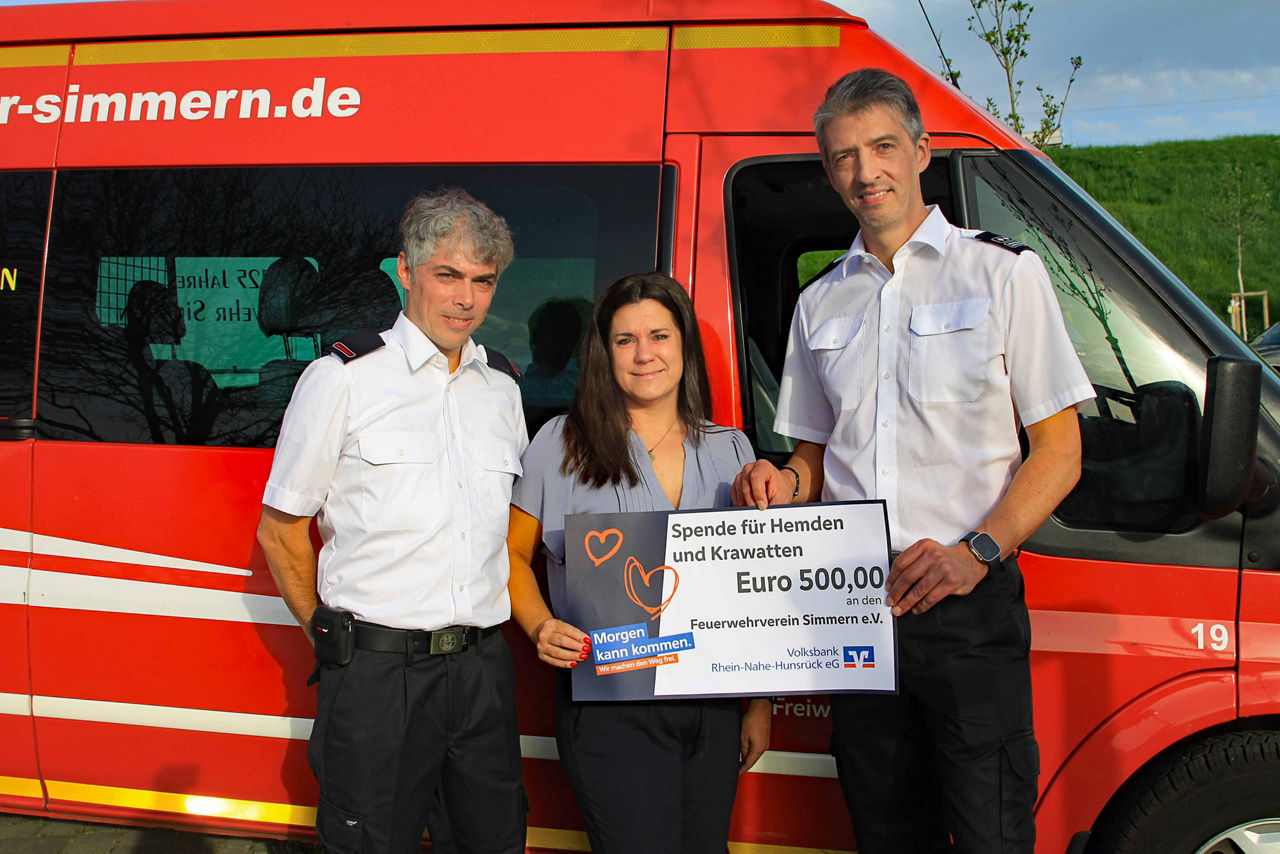 Drei Erwachsene stehen vor einem Feuerwehrauto und halten einen Spendenscheck in der Hand.