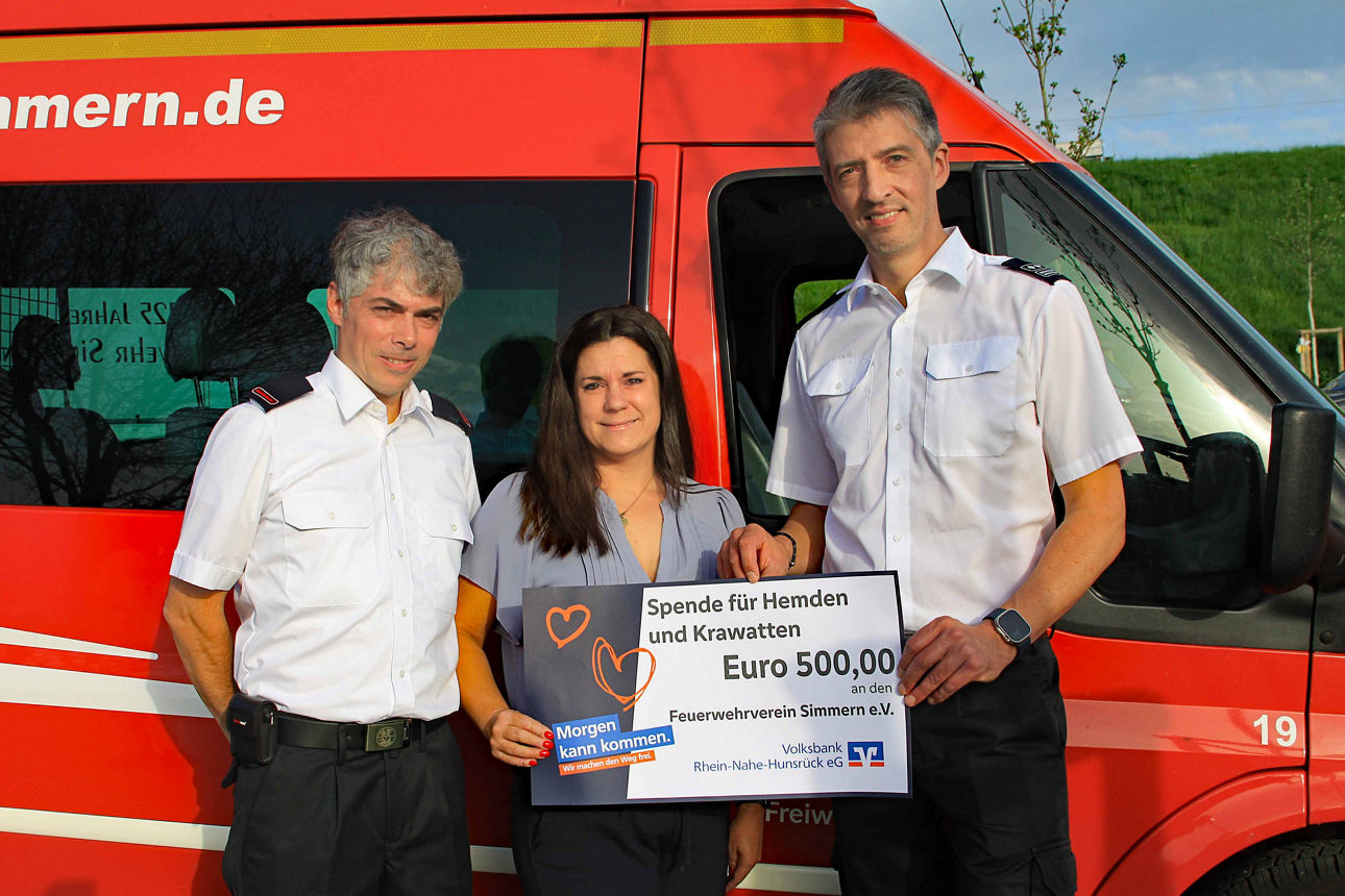 Drei Erwachsene stehen vor einem Feuerwehrauto und halten einen Spendenscheck in der Hand.