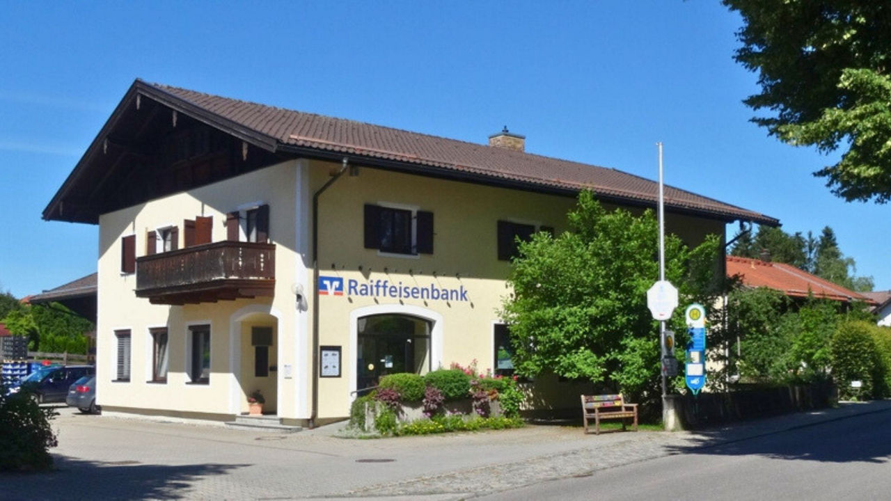 Eine traditionelle Bankfiliale der Raiffeisenbank in Dietramszell, einem zweistöckigen Gebäude mit gelber Fassade, Holzbalkon und einem Schild mit dem Raiffeisenbank-Logo. Die Filiale ist von Grünflächen und Bäumen umgeben.