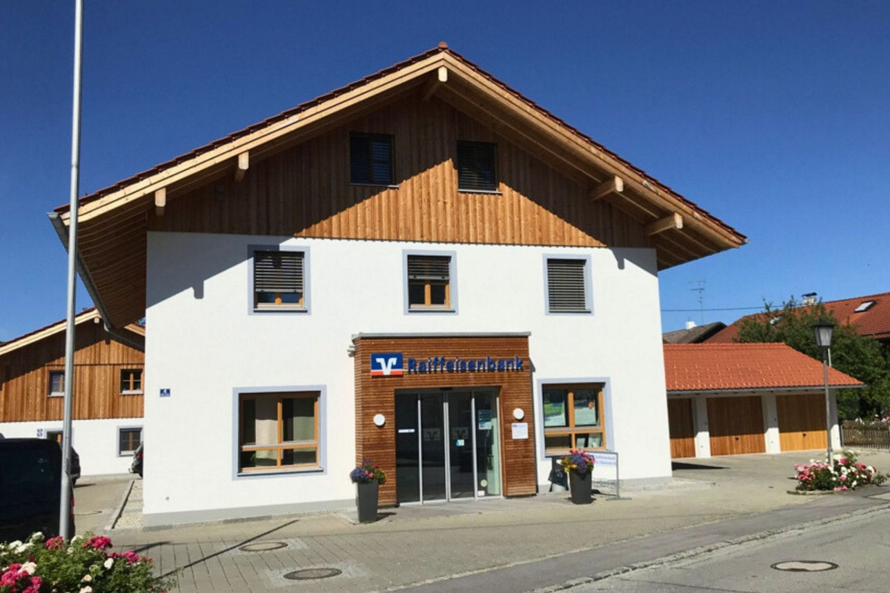 Eine moderne Bankfiliale der Raiffeisenbank in Egling, einem zweistöckigen Gebäude mit weißer Fassade und Holzgiebel. Der Eingangsbereich ist von Blumenkübeln gesäumt.