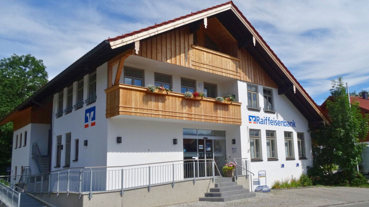 Eine moderne Bankfiliale der Raiffeisenbank in Gaißach, einem zweistöckigen Gebäude mit weißer Fassade, Holzgiebel und Balkon. Das Gebäude verfügt über große Fenster und einen barrierefreien Eingang. Blumenkästen mit roten und weißen Blumen schmücken den Balkon.