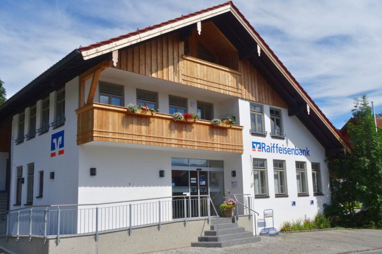 Eine moderne Bankfiliale der Raiffeisenbank in Gaißach, einem zweistöckigen Gebäude mit weißer Fassade, Holzgiebel und Balkon. Das Gebäude verfügt über große Fenster und einen barrierefreien Eingang. Blumenkästen mit roten und weißen Blumen schmücken den Balkon.