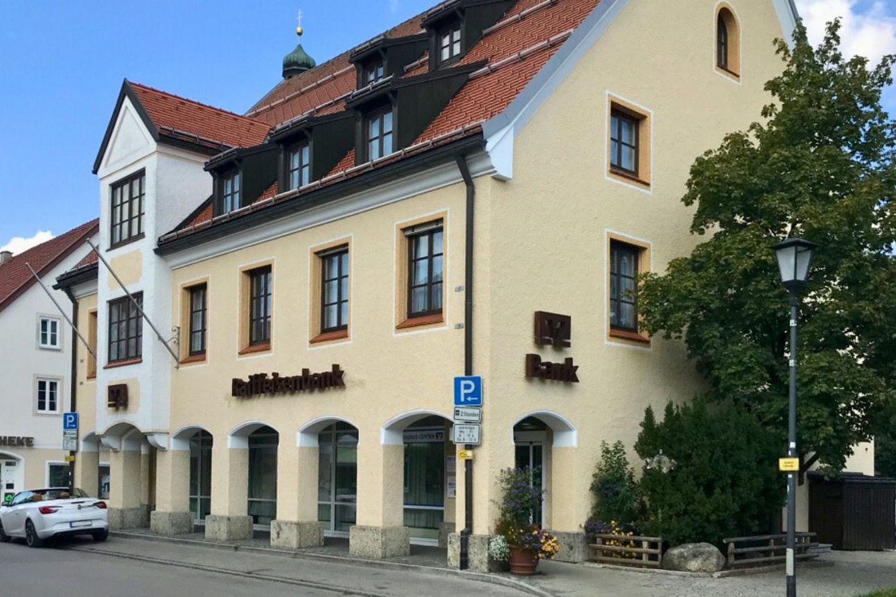 Eine traditionell gestaltete Bankfiliale der Raiffeisenbank in Lenggries, einem mehrstöckigen Gebäude mit heller Fassade, roten Ziegeldächern und markanten Arkaden im Erdgeschoss. Ein weißes Auto ist vor dem Gebäude geparkt.