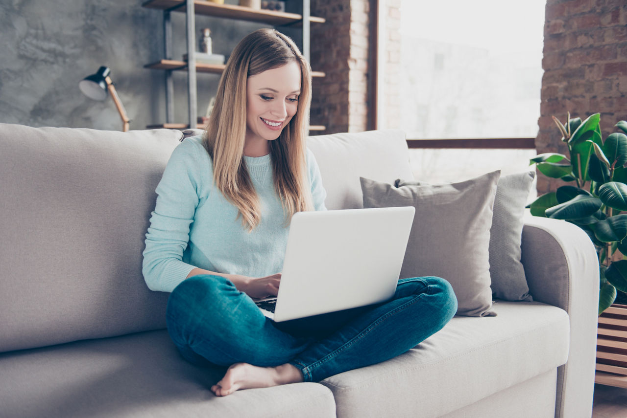 Porträt eines positiven, hübschen, charmanten, häuslichen Mädchens, das auf dem Sofa sitzt und einen Laptop auf ihren Beinen 