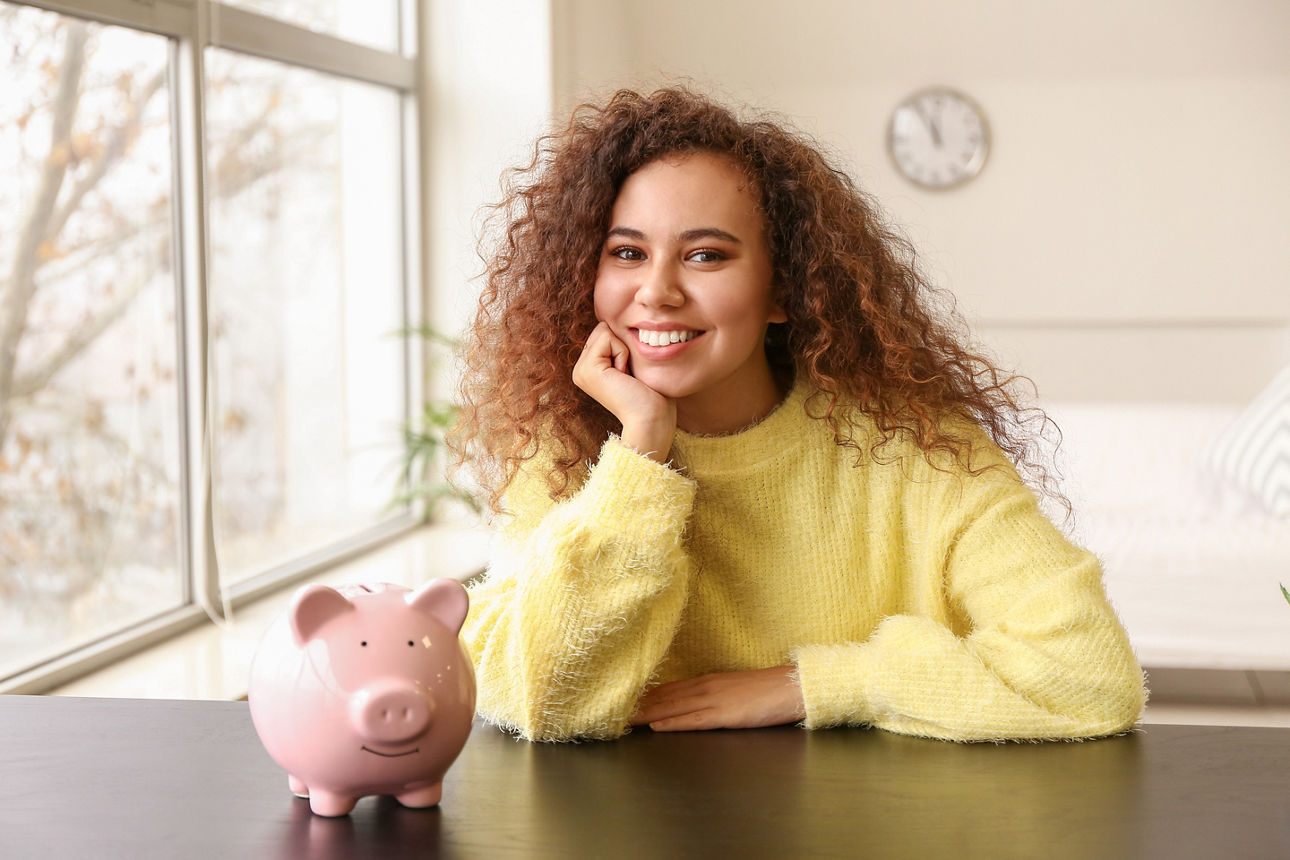 Eine Frau sitzt an einem Tisch, auf dem ein rosanes Sparschwein steht.