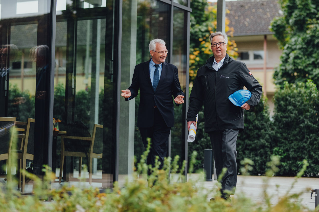 Ein Mitarbeiter führt ein Gespräch mit einem Kunden, währenddessen laufen sie an einem Gebäude vorbei