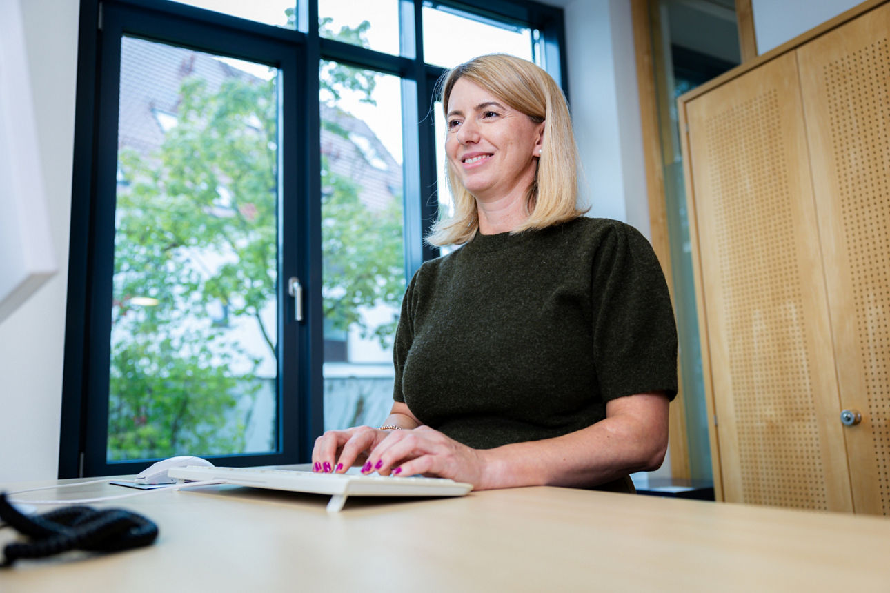 Mann im Anzug sitzt lächelnd am Arbeitsplatz vor Monitor und Fenster.