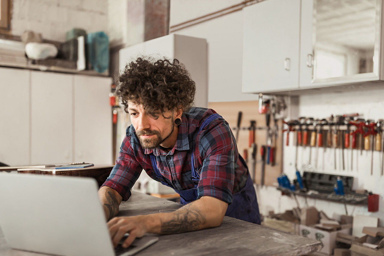 In einer Schreinerwerkstatt beugt sich der Schreiner an einem Tisch über den aufgeklappten Laptop vor sich. Der Schreiner hat wilde Locken, einen Bart, tatowierte Arme und ein hochgekrempeltes Karohemd an unter seinem Blaumann. Allgemein würde man ihn als modern beschreiben. 