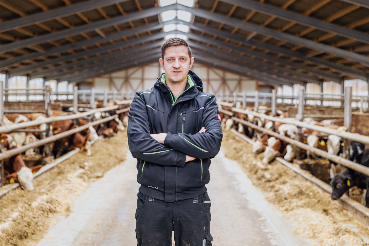 Markus Fischer steht im Mittelgang eines Tierwohl‑Stalls, flankiert von Rindergruppen an den Futterplätzen. Sichtbar sind die Stallstruktur, Fressgitter sowie Stroheinstreu im Fütterungsbereich des Stalls.