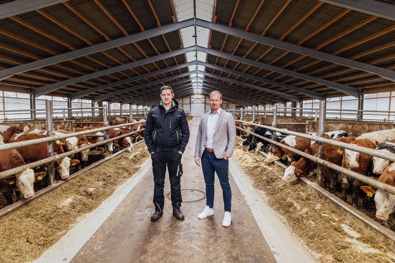 Markus Fischer und Berater Florian Abschütz befinden sich im Innenbereich eines Tierwohl‑Stalls mit Strohhaltung. Links und rechts sind Rinder an den Futterplätzen zu sehen, während im Mittelgang der Stallstruktur der Produktions‑ und Arbeitsbereich erkennbar ist