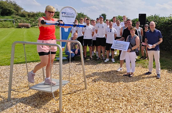 Eine Frau mittleren Alters testet ein Outdoor-Fitnessgerät mit vielen lächelnden Menschen im Hintergrund