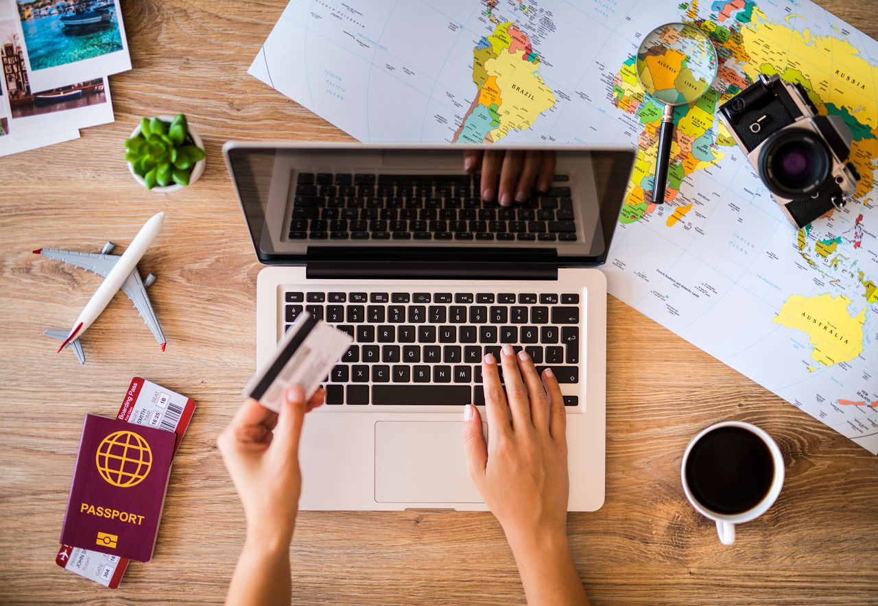 Hände buchen Reise online am Laptop mit Karte, Weltkarte, Kamera und Pass auf dem Tisch.