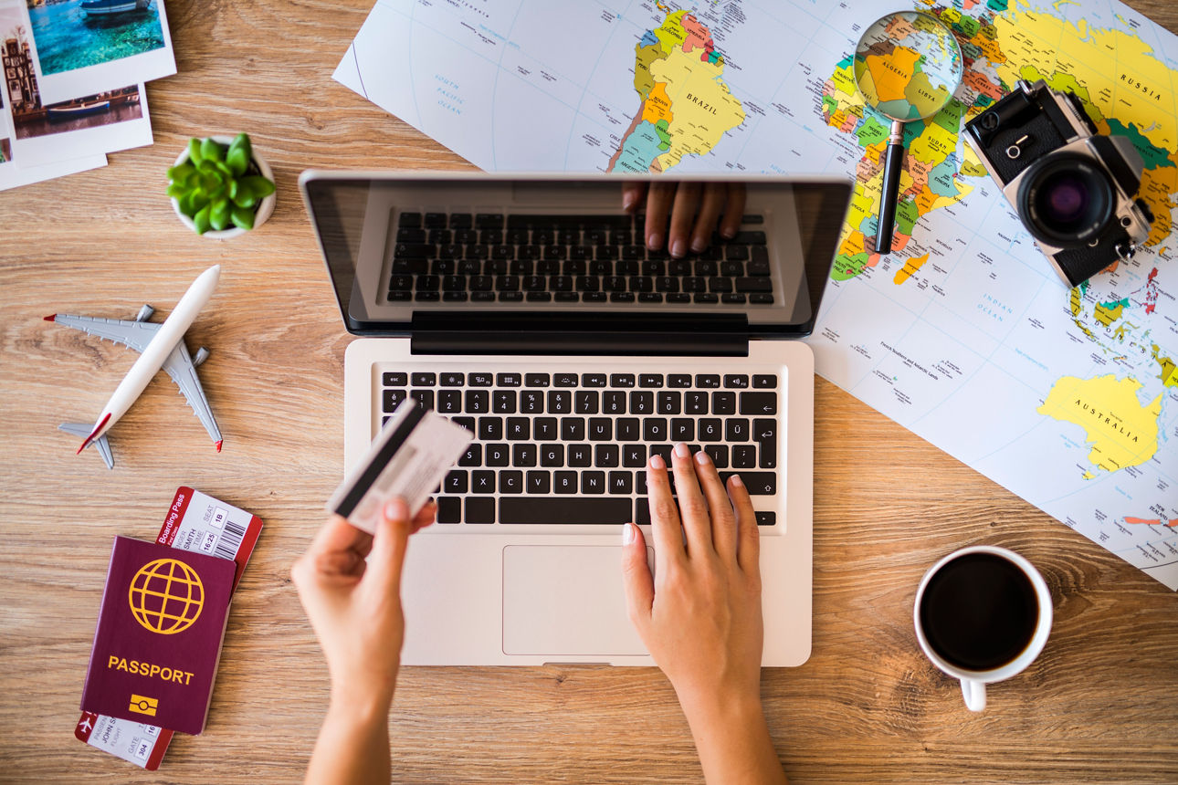 Hände buchen Reise online am Laptop mit Karte, Weltkarte, Kamera und Pass auf dem Tisch.