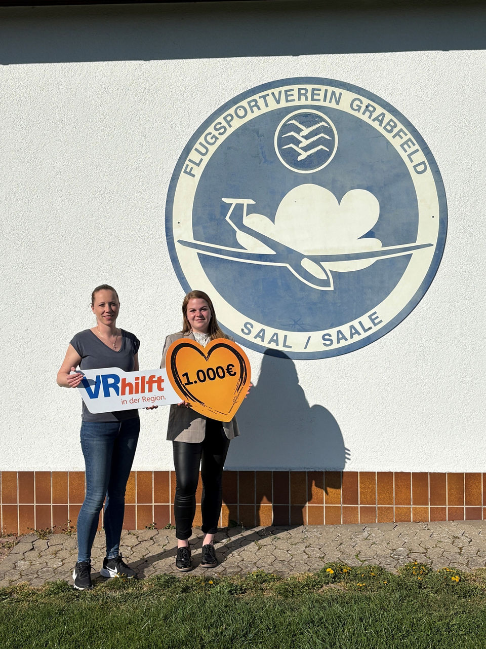 Das Bild zeigt zwei Frauen, die vor einer weißen Wand mit einem Logo stehen. Auf dem Logo steht: Flugsportverein Grabfeld. Si