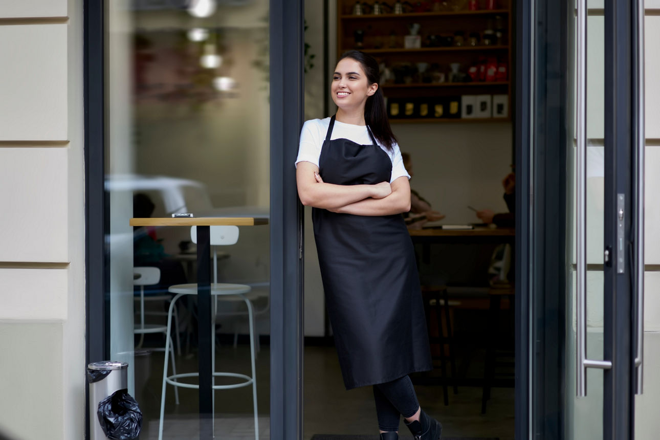 Eine Frau steht in der Ladentür zu ihrem Geschäft und lächelt, während sie sich umschaut.