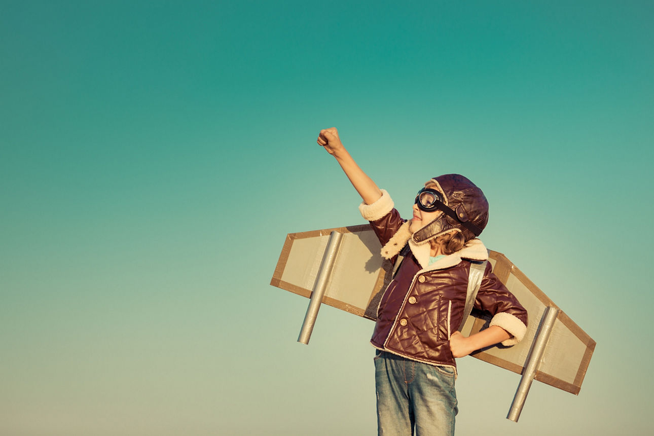 Ein Kind, das eine Fliegerjacke, eine Fliegerhaube und Schutzbrille trägt, mit aufgesetzten Pappflügeln