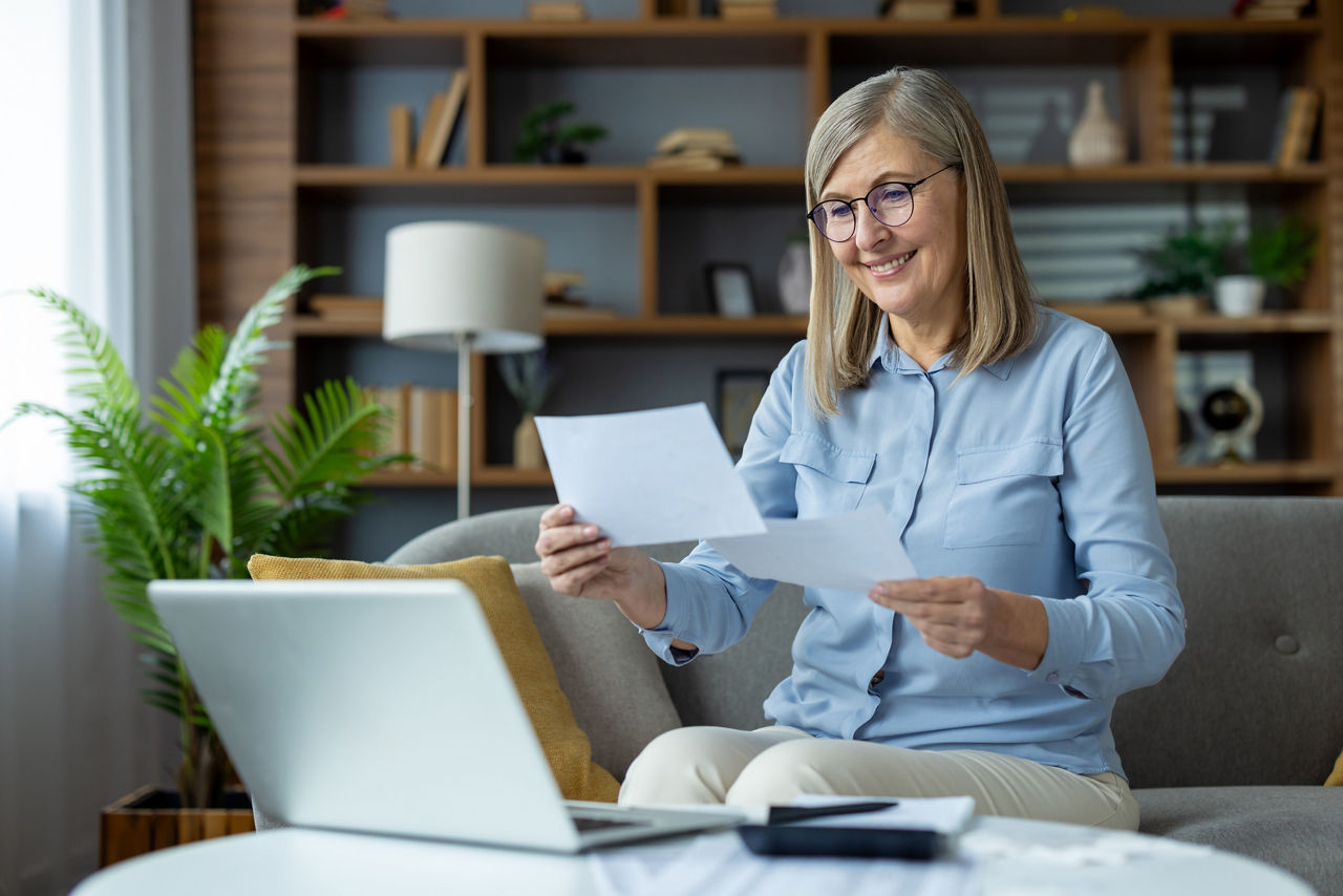 Ältere Frau sitzt auf einem Sofa und hält in jeder Hand ein Blatt Papier. Vor ihr ist eine Kaffeetisch mit einem Laptop zu sehen